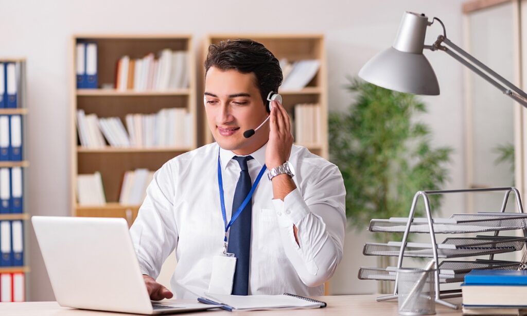 a man sitting at a laptop while holding the headset on his ear