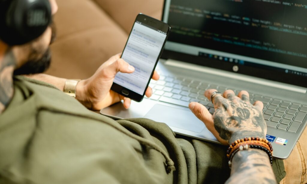 man with many tattoos holding a phone and laptop looks at the screens