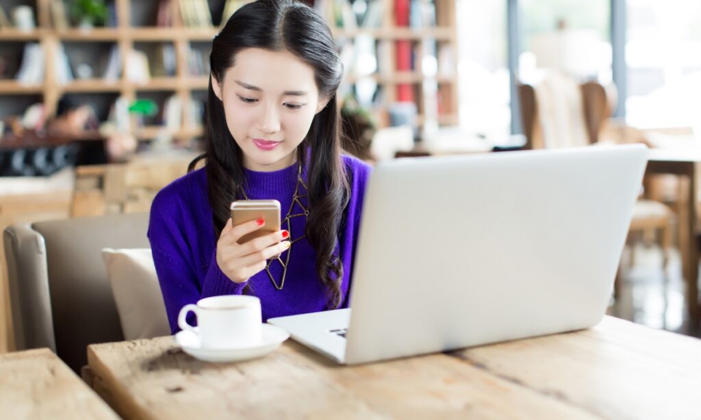 a young woman in a cafe reading on her phone with a laptop and coffee cup