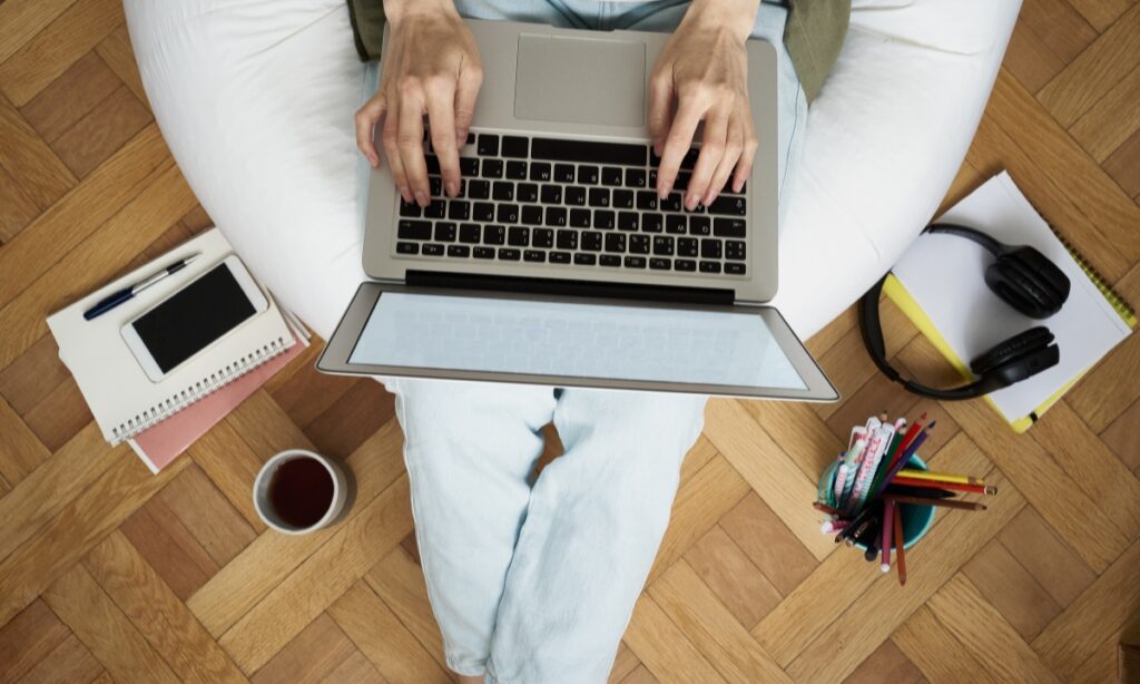 a person sits in a beanbag chair and holds a laptop with work items around them, like notebooks, pens, and coffee