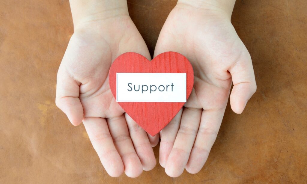 a person's hands holding a cardboard heart that says  "support" 