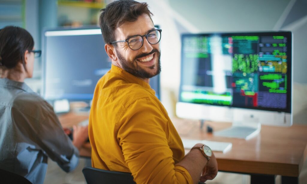 man wearing yellow shirt and glasses smiles over his shoulder in front of a computer