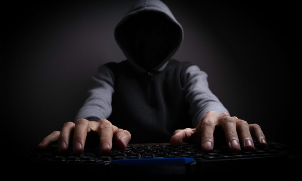 a hacker with their face covered by a dark hood with their hands on a keyboard