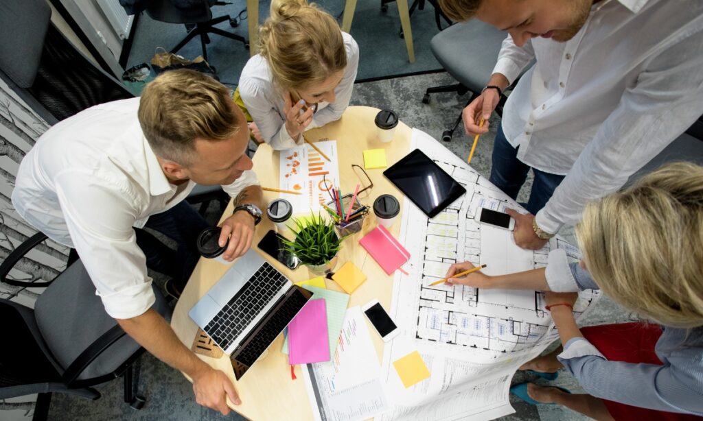 a group of people around a bunch of papers, files, laptops, and tablets