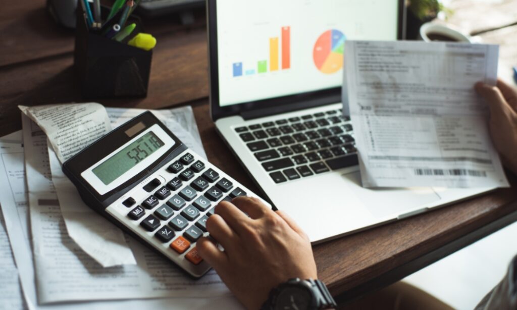 a person looking at paperwork and using a laptop and calculator to calculate costs