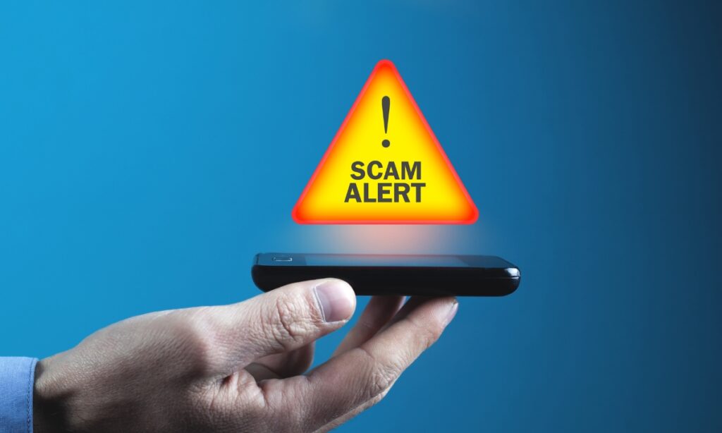 a person's hand holding an old cell phone with a red and orange triangle floating above it that says "scam alert"