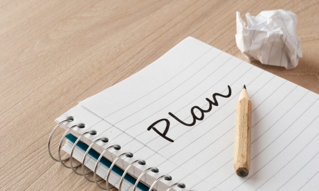 a notebook with a wooden pencil and lined paper that says "plan." There is a crumpled piece of paper next to the notebook