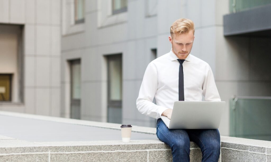 a man sitting on the edge of a building with a laptop on his lap and a coffee next to him